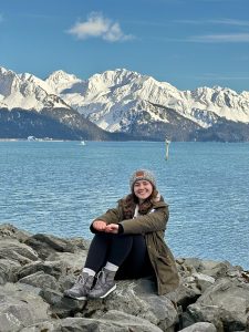 sarah markovich sitting on rocky area in front of blue water and snowy mountains