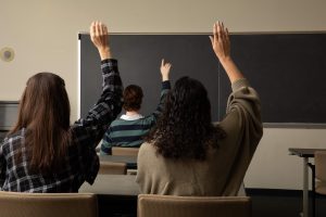 The backs of three female high school students sitting at their desks and raising their hands