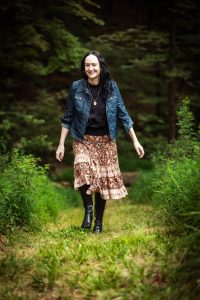 woman with dark hair in a denim jacket, long patterned skirt and boots walking through the woods