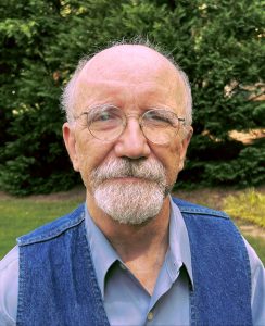 photo of man in round, wire-rimmed glasses wearing a blue button-down shirt and blue vest