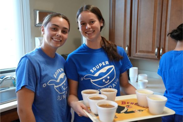 Two smiling students in "Souper Sunday" t-shirts hold a tray of soup and prepare to serve it