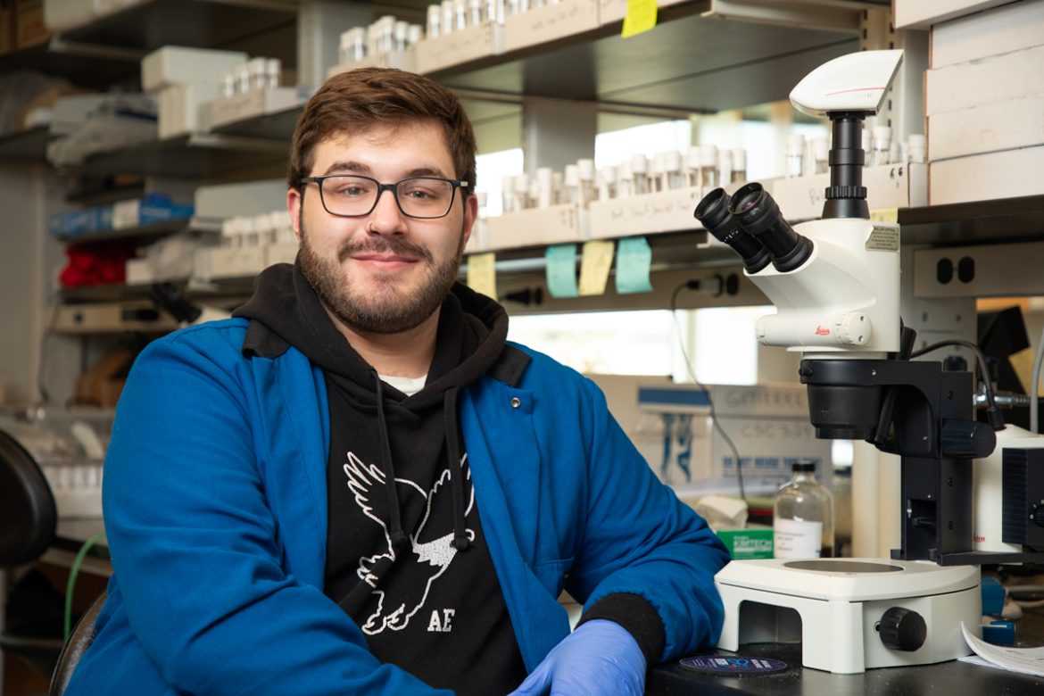 college student wearing a lab coat seated by a microscope